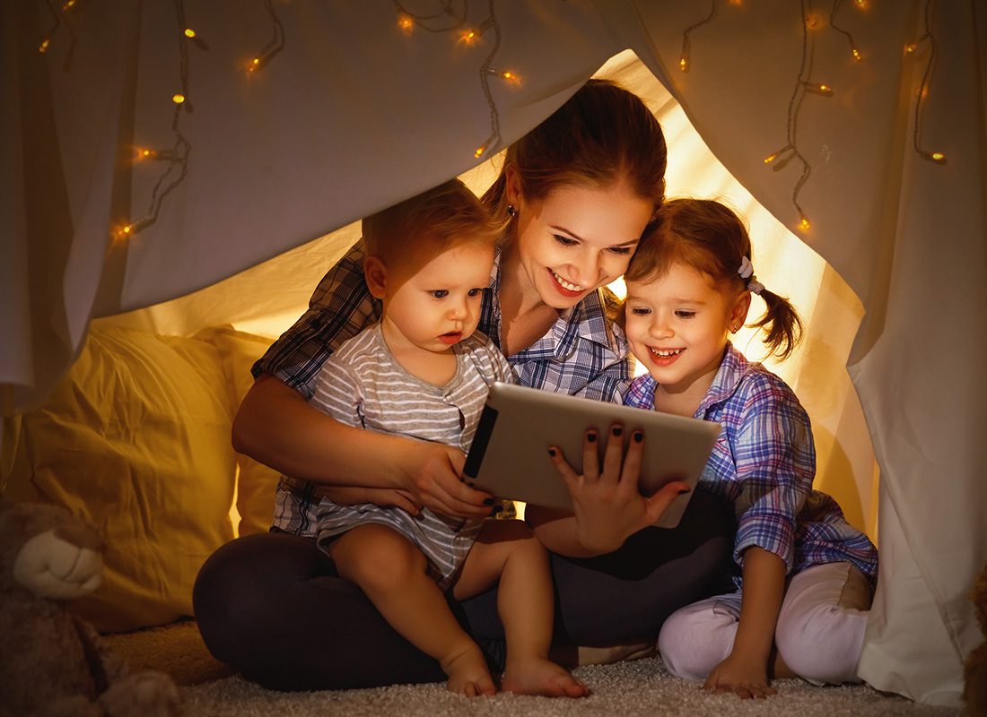 Blog - Mother Sitting on Floor with Two Daughters Under a Tent Made of Sheets and Twinkeling Lights While They Look at a Tablet Smiling